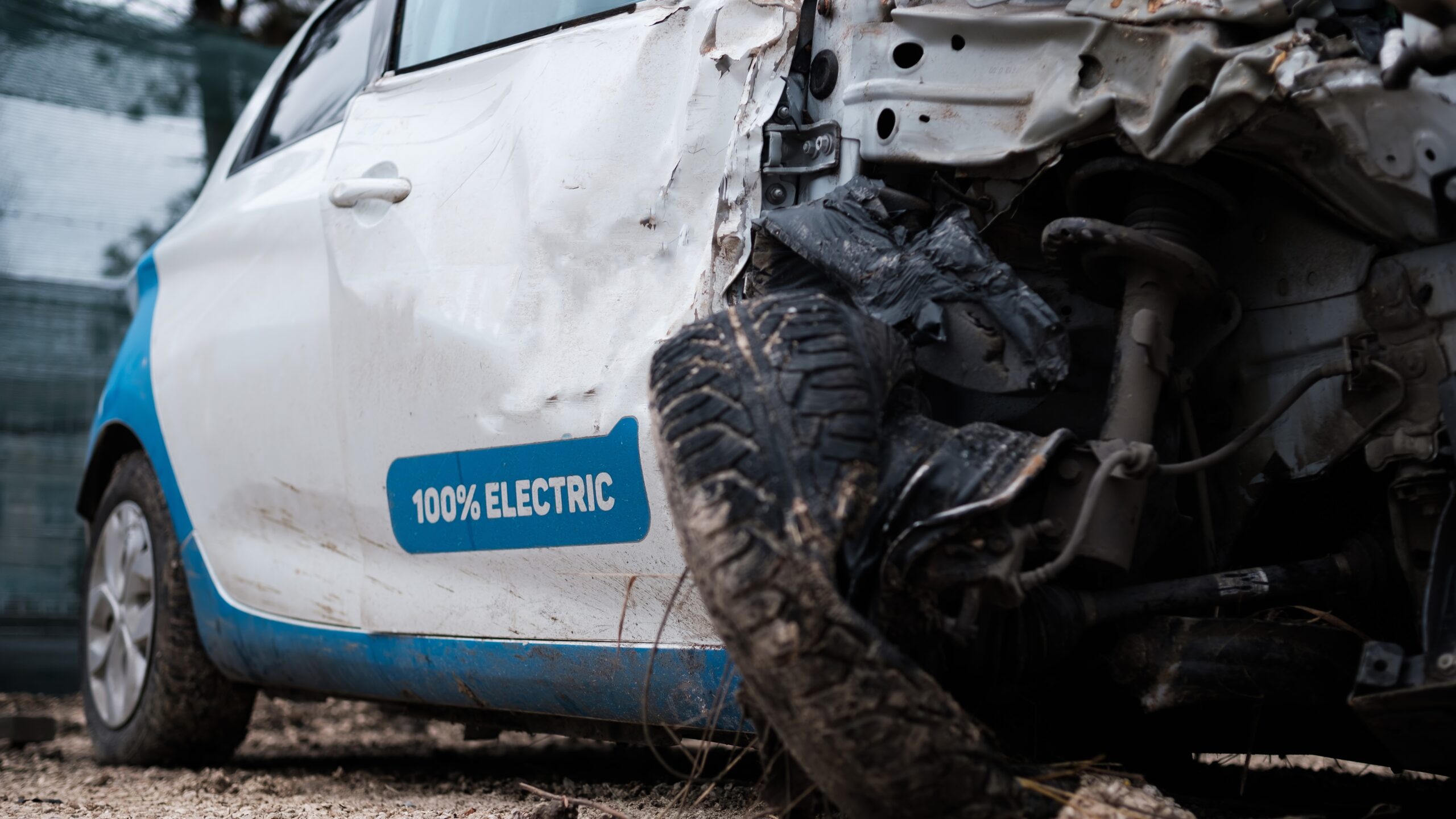 Ein kleines weißes Elektroauto mit schweren Frontschäden und verbeulter Motorhaube steht mit offener Vordertür in der Nähe eines Maschendrahtzauns. Das Auto mit der Aufschrift „100 % ELEKTRISCH“ benötigt aufgrund des Schadensausmaßes wahrscheinlich eine Scheinwerferreparatur.